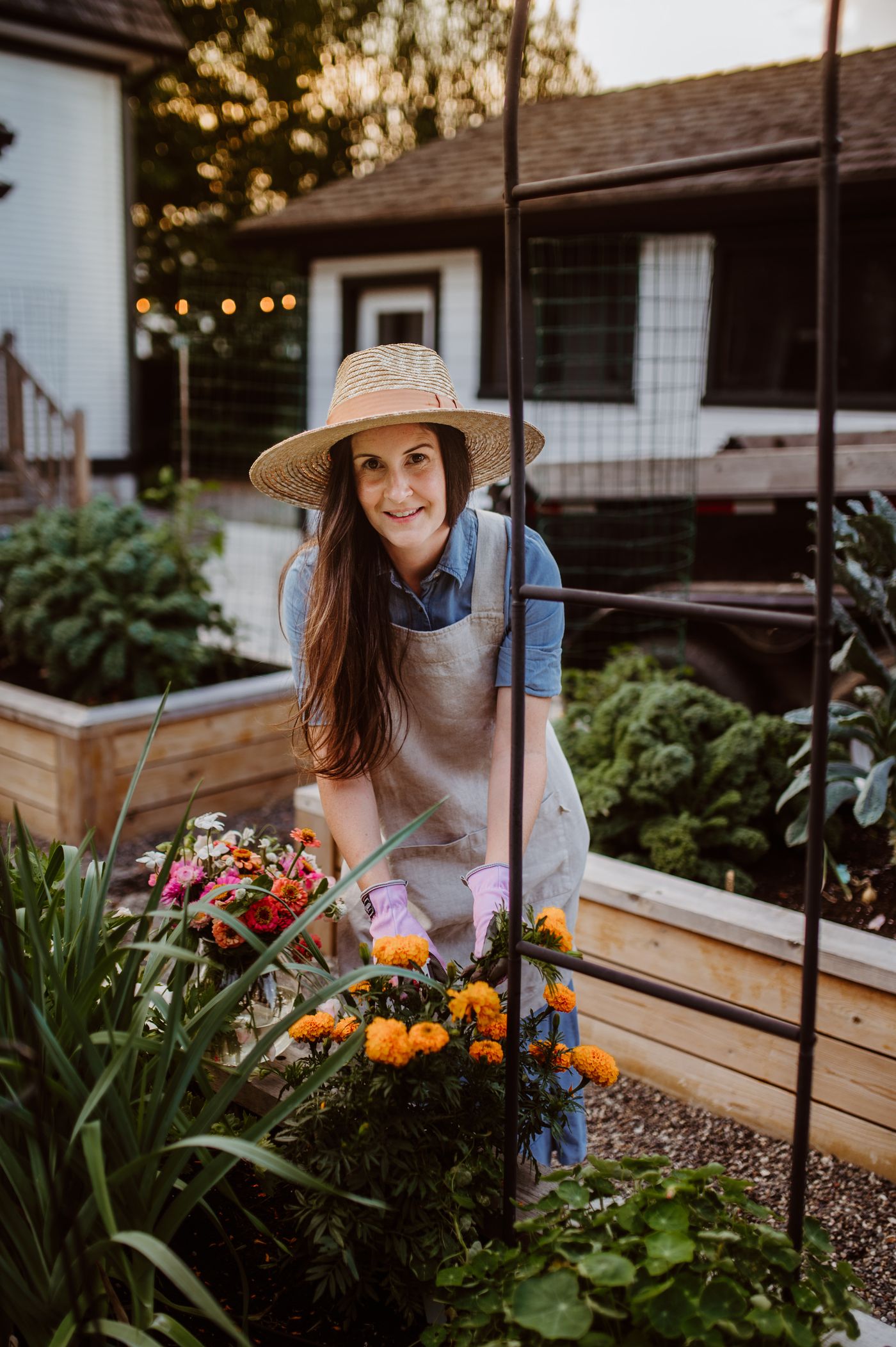 A gardener in a sun hat harvesting from raised garden beds in a backyard at golden hour